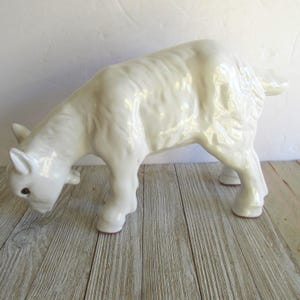 May include: A white ceramic goat figurine with a downward gaze. The goat has a textured coat and is posed with its head lowered. The hooves have a reddish-brown color. The figurine is on a wooden surface.