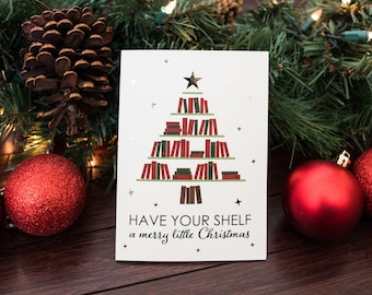 May include: A white Christmas card with a gold star and a green Christmas tree made of books. The text on the card reads "HAVE YOUR SHELF a merry little Christmas".