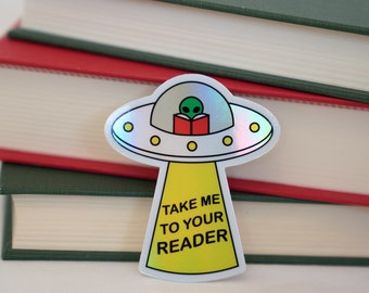 Puede incluir: Una pegatina holográfica de un OVNI con un extraterrestre leyendo un libro. El OVNI tiene un rayo de luz amarillo que brilla hacia abajo y el texto "Take me to your reader" está escrito en el rayo.