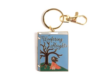 May include: Gold keychain featuring a blue enamel book with the title "Wuthering Heights" and a black silhouette of a tree with a woman in a red dress standing beneath it.