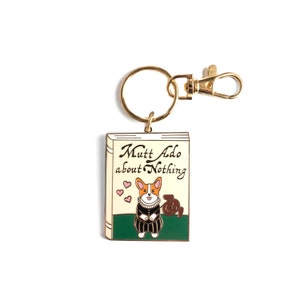 May include: Gold-tone keychain featuring a book-shaped charm with a cartoon corgi wearing a black and white dress and holding a brown dog. The book charm has the text "Mutt Ado about Nothing" on the cover.