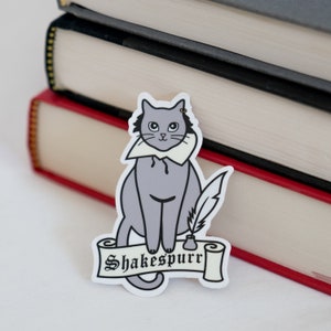 Puede incluir: Un gato gris con un collar blanco y sosteniendo una pluma de ave se sienta en un rollo con el texto "Shakespurr". El gato es una pegatina con fondo blanco.