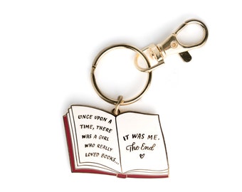 Puede incluir: Llavero dorado en forma de libro con borde rojo. El llavero presenta el texto "ONCE UPON A TIME, THERE WAS A GIRL WHO REALLY LOVED BOOKS... IT WAS ME. The End." con un símbolo de corazón.
