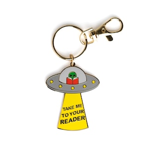 Pode incluir: Um chaveiro dourado com um amuleto de UFO cinza e amarelo. O UFO tem um alienígena verde dentro lendo um livro vermelho. O amuleto diz "Leve-me ao seu leitor".