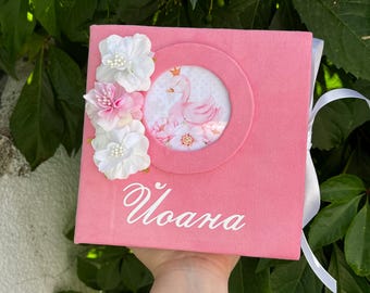 May include: Pink photo album with a circular image of a swan wearing a crown, surrounded by flowers. The album is decorated with white and pink fabric flowers and the name "Ioana" in cursive.