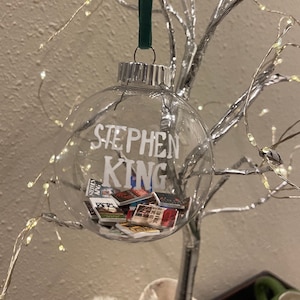 May include: A clear glass Christmas bauble with the name "Stephen King" written on it. Inside the bauble are miniature versions of Stephen King books.