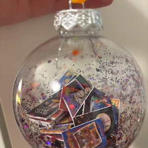 May include: A clear spherical Christmas bauble filled with purple and black glitter and miniature book covers. The bauble features a silver metal cap with a yellow hanging loop. The miniature book covers include titles like Hocus Pocus and Goosebumps.