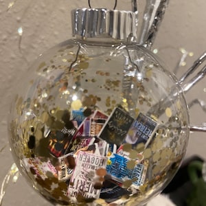 May include: A clear glass Christmas bauble filled with gold glitter and miniature book covers. The bauble has a silver top and a green ribbon.