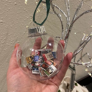 May include: A clear glass Christmas bauble filled with miniature book covers featuring Stephen King novels. The bauble is hanging from a green ribbon.