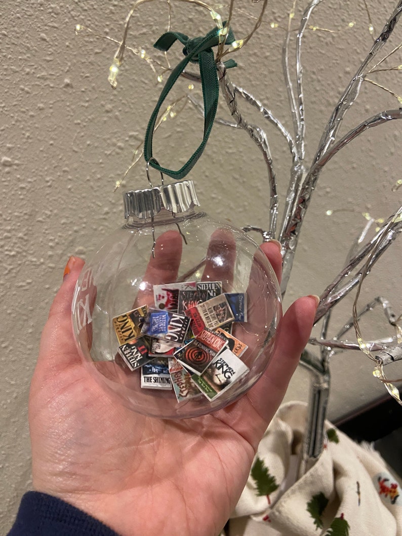 May include: A clear glass Christmas bauble filled with miniature book covers featuring Stephen King novels. The bauble is hanging from a green ribbon.