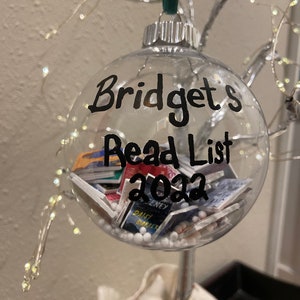 May include: A clear glass bauble filled with white beads. The bauble is decorated with black marker writing that says "Bridget's Read List 2022".