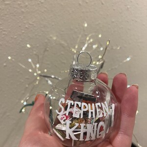 May include: A clear glass Christmas bauble with a silver top and a silver wire loop. The bauble is filled with miniature book covers and the words "Stephen King" are written on the front.