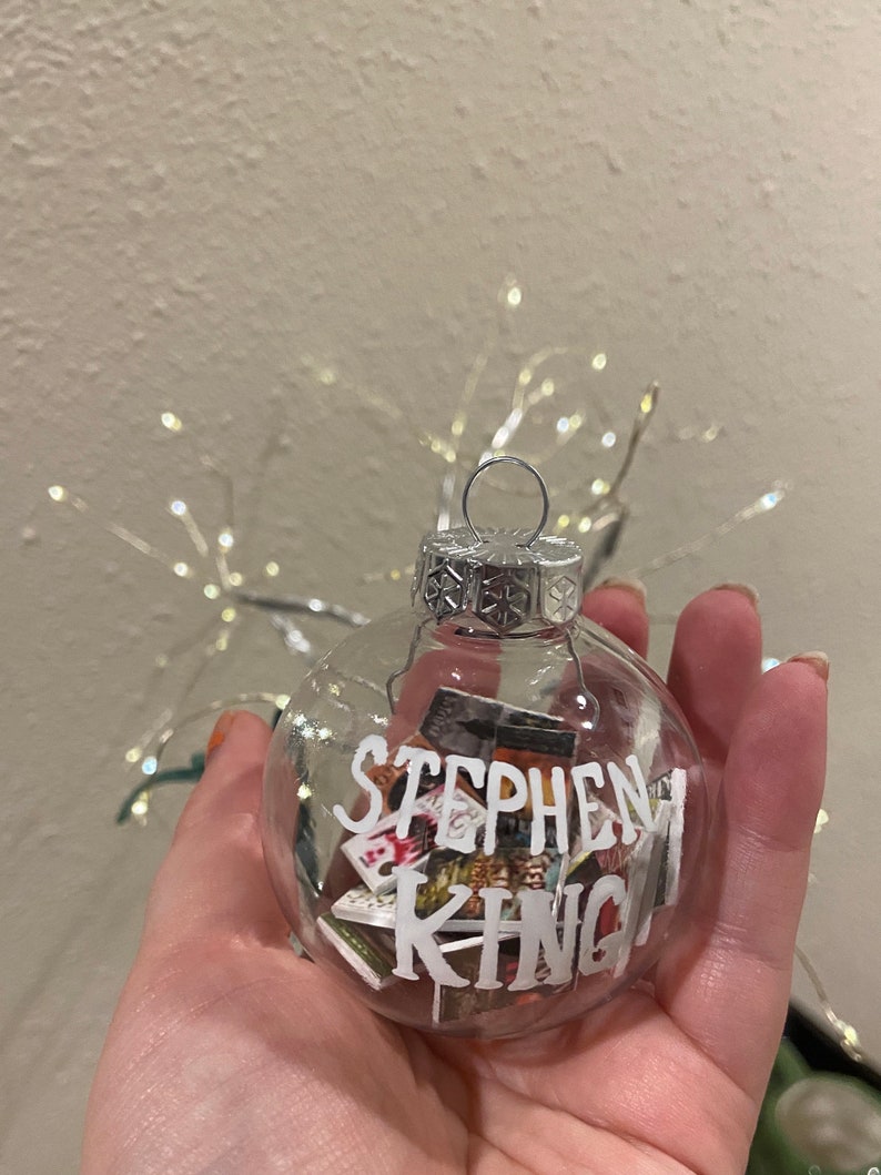 May include: A clear glass Christmas bauble with a silver top and a silver wire loop. The bauble is filled with miniature book covers and the words "Stephen King" are written on the front.