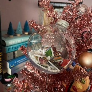 May include: A clear glass Christmas ornament filled with white beads and small colorful cards. The ornament is hanging on a pink tinsel Christmas tree.