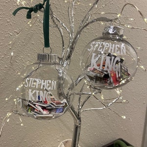May include: Two clear glass baubles with silver tops and white lettering that reads "Stephen King". The baubles are filled with small, colourful book covers and are hanging from a silver wire tree.