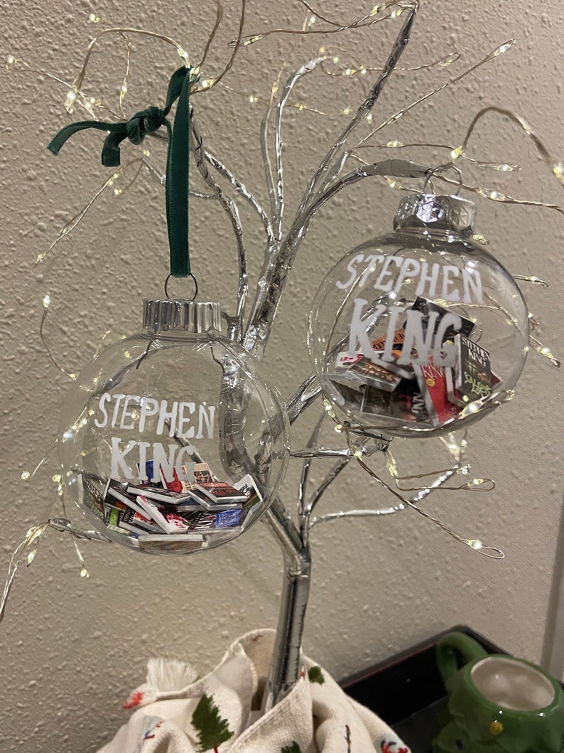 May include: Two clear glass baubles with silver tops and white lettering that reads "Stephen King". The baubles are filled with small, colourful book covers and are hanging from a silver wire tree.