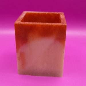 Puede incluir: Un pequeño macetero cuadrado hecho de resina blanca y naranja. El macetero tiene una superficie lisa, ligeramente texturizada y un sutil efecto degradado.