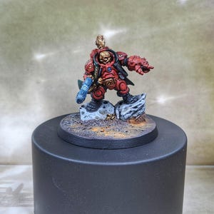 Könnte beinhalten: Eine rot und gold bemalte Miniaturfigur eines Space Marines, der eine blaue Waffe hält. Die Figur steht auf einem grauen und braunen Sockel.