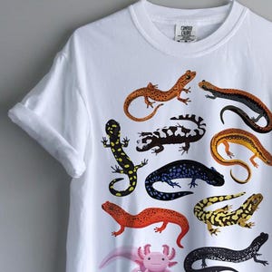 May include: White t-shirt featuring colorful illustrations of various salamanders. The designs include salamanders in shades of orange, black, yellow, and blue. The shirt is hanging on a white hanger against a gray wall.