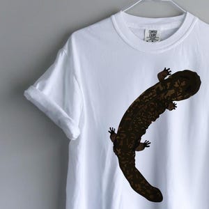 May include: A white short-sleeved t-shirt featuring a large, dark brown, mottled salamander graphic centered on the chest. The shirt has a crew neckline and rolled sleeves, displayed on a white wire hanger against a plain light gray background.
