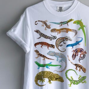 以下が含まれることがあります： 白の半袖Tシャツ。前面にはヤモリやカメレオンなど、様々な種類のトカゲがグリッド状に配置されたカラフルなグラフィックプリントが施されています。クルーネックで、袖口はロールアップされたデザインです。