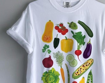 May include: A white t-shirt featuring a colourful graphic print of various vegetables including a butternut squash, tomatoes, a yellow bell pepper, an aubergine, a red onion, a carrot, corn, lettuce, peas, and cucumber slices arranged in a central cluster.