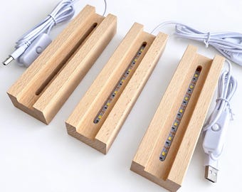 Puede incluir: Tres bases de exhibición rectangulares de madera clara, cada una con una ranura central. Dos de las bases contienen tiras de luz LED integradas. Cada base está conectada a un cable de alimentación USB blanco con un interruptor integrado.