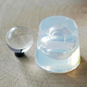 Könnte beinhalten: Eine klare Glaskugel und eine klare Silikonform zum Gießen von Harz. Die Form ist wie ein kleiner, runder Behälter mit einem erhabenen, kreisförmigen Design in der Mitte geformt.
