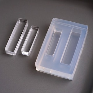 Pode incluir: Dois moldes de silicone retangulares transparentes para fazer artesanato de resina. O molde maior tem duas cavidades retangulares, enquanto o molde menor tem uma cavidade retangular.