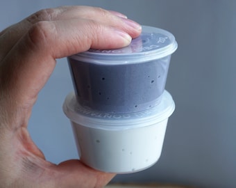 Puede incluir: Dos pequeños recipientes de plástico de slime, uno morado y otro blanco, apilados uno encima del otro. Los recipientes tienen tapas transparentes.