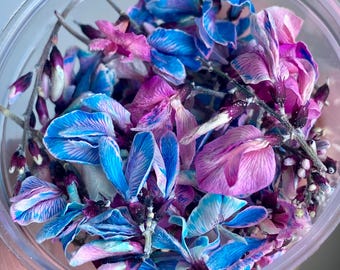 Puede incluir: Un recipiente de plástico transparente lleno de pétalos de flores azules y moradas secas. Los pétalos tienen una apariencia delicada y en capas, con tonos de azul, morado y rosa. El recipiente se sostiene en una mano, con el fondo desenfocado.
