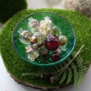 May include: A green bowl filled with small glass ornaments. The ornaments are filled with dried flowers and other botanicals. Some of the ornaments are clear, while others are a deep red color.