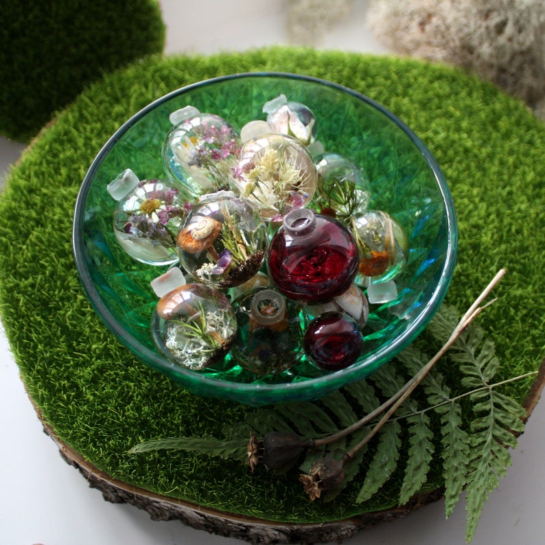 May include: A green bowl filled with small glass ornaments. The ornaments are filled with dried flowers and other botanicals. Some of the ornaments are clear, while others are a deep red color.
