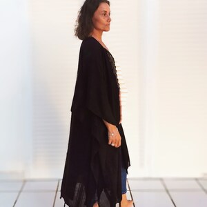 K&ouml;nnte beinhalten: Eine Frau tr&auml;gt ein schwarzes, lang&auml;rmliges, flie&szlig;endes Kimono mit Fransen am Saum. Sie steht auf einem wei&szlig;en Fliesenboden.