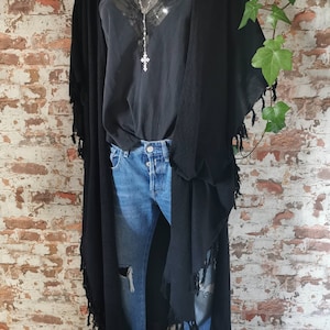 K&ouml;nnte beinhalten: Ein schwarzer Langarm-Cardigan mit Fransenbesatz. Der Cardigan wird &uuml;ber einem schwarzen Tanktop mit Spitzenbesatz und einer silbernen Kettenkette mit einem Kreuzanh&auml;nger getragen. Die Person tr&auml;gt eine blaue Jeans.