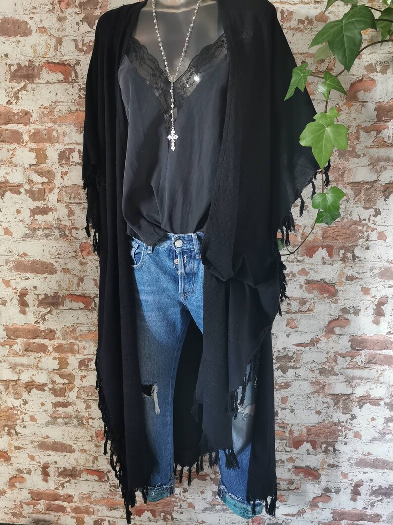 K&ouml;nnte beinhalten: Ein schwarzer Langarm-Cardigan mit Fransenbesatz. Der Cardigan wird &uuml;ber einem schwarzen Tanktop mit Spitzenbesatz und einer silbernen Kettenkette mit einem Kreuzanh&auml;nger getragen. Die Person tr&auml;gt eine blaue Jeans.