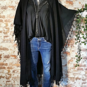 K&ouml;nnte beinhalten: Schwarzer Kimono mit langen &Auml;rmeln und Fransen. Der Kimono wird &uuml;ber einem schwarzen Tanktop und einer blauen Jeans getragen.