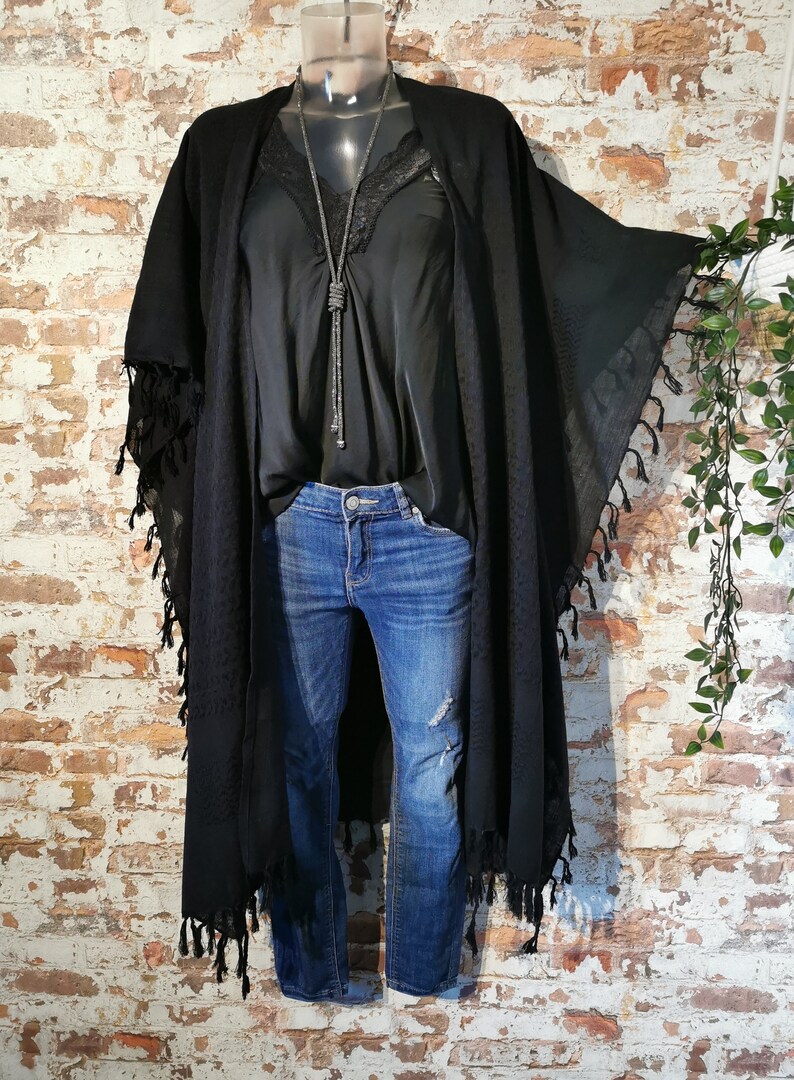 K&ouml;nnte beinhalten: Schwarzer Kimono mit langen &Auml;rmeln und Fransen. Der Kimono wird &uuml;ber einem schwarzen Tanktop und einer blauen Jeans getragen.