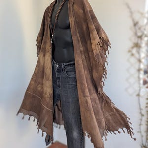 Könnte beinhalten: Ein brauner Kapuzenponcho mit Quastenbesatz ist über einer schwarzen Schaufensterpuppe drapiert. Der Poncho wird mit dunkelblauen Jeans und einer Perlenkette kombiniert. Das Kleidungsstück hat einen Batik-Effekt und hängt vor einer weißen Wand.