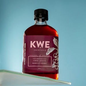 Puede incluir: Una botella de vidrio de jarabe de cereza para cócteles. La etiqueta es roja y presenta el texto "KWE Cocktails", "Recolte Unique", "Single Harvest", "Sirop Cocktails Syrup", "Cerise Griotte", "Morello Cherry" y "200 ml".