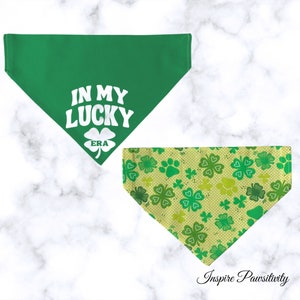 May include: Two green dog bandanas. One bandana is solid green with the text "In My Lucky Era" in white. The other bandana has a pattern of green four-leaf clovers and paw prints on a light yellow background.