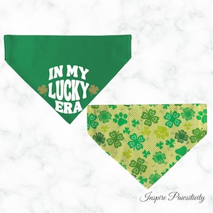 May include: Two dog bandanas. One is solid green with the text "IN MY LUCKY ERA" and shamrock accents. The other is patterned with shamrocks and paw prints in shades of green on a yellow background. The bandanas are triangular.