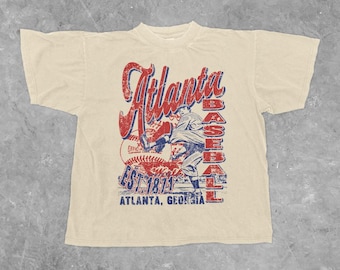 Può includere: T-shirt color crema con un design grafico invecchiato. Il design include la parola "Atlanta" in rosso, le parole "BASEBALL" in rosso e blu e il testo "EST 1871 ATLANTA, GEORGIA". La grafica raffigura un giocatore di baseball.