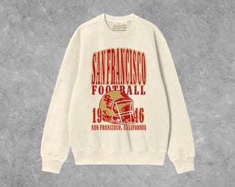 Op de afbeelding: Crèmekleurige sweatshirt met een rode en gouden afbeelding. De afbeelding leest "SAN FRANCISCO FOOTBALL 1946" met een illustratie van een voetbalhelm. De tekst "SAN FRANCISCO, CALIFORNIA" staat eronder.