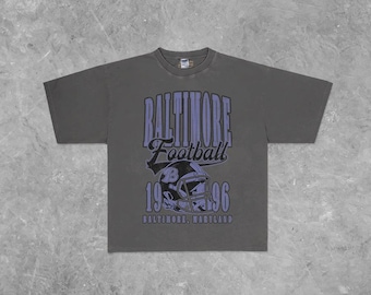 Puede incluir: Camiseta gris oscuro con la inscripción "BALTIMORE Football" en morado, con una fuente de estilo vintage. Debajo, un gráfico de casco de fútbol morado, el número "1996" y el texto "BALTIMORE, MARYLAND".