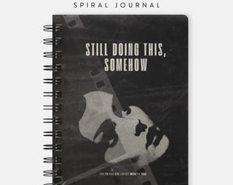 May include: A black spiral-bound journal from the SALTMOTH COLLECTION. The cover features a film strip graphic, a white abstract shape, and the text: STILL DOING THIS, SOMEHOW and TIME TO MAKE EYE CONTACT WITH THE VOID.