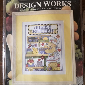 Puede incluir: Un kit de punto de cruz contado con un oso de dibujos animados en una cocina. El kit incluye tela Aida de algodón 100%, hilo de bordar, una aguja e instrucciones ilustradas. El kit se titula "Julie's Kitchen".