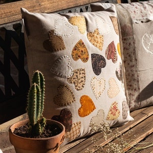 Puede incluir: Un cojín decorativo con fondo beige y varios parches en forma de corazón en tonos marrón, amarillo y naranja. El cojín está colocado en un banco de madera junto a un cactus en maceta. Otro cojín con diseño de corazón es parcialmente visible.