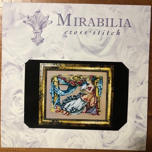 Può includere: Uno schema di punto croce con una donna in un abito blu e oro che tiene un piccione bianco. Lo schema è incorniciato in una cornice dorata con un passepartout nero. Lo sfondo è un motivo floreale bianco.
