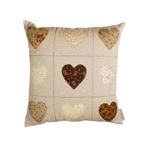 Puede incluir: Una almohada beige con un diseño de patchwork de nueve corazones en varios tonos de marrón, beige y blanco. Los corazones están hechos de diferentes telas con diseños florales y estampados.